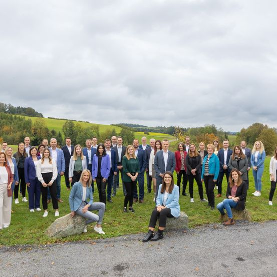 Eine große Gruppe von Geschäftsleuten, gekleidet in Geschäftskleidung, posiert für ein Foto auf einer Wiese, mit einem Schotterweg und grünen Hügeln im Hintergrund.