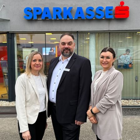 Drei Personen stehen vor einer Bank und lächeln für ein Foto. Der Mann steht in der Mitte, und zwei Frauen sind auf beiden Seiten. Sie stehen vor einer Bankfiliale.