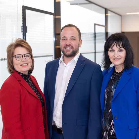 Drei Personen stehen nebeneinander, lächelnd, in einem Büro mit Glaswänden.