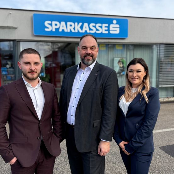 Drei Personen, gekleidet in formeller Kleidung, stehen vor einem Gebäude mit einem blauen Schild, auf dem 'SPARKASSE' steht. Der Mann auf der linken Seite hat die Hände in den Taschen, der Mann in der Mitte hat einen Bart und lächelt, und die Frau auf der rechten Seite trägt eine Brille.