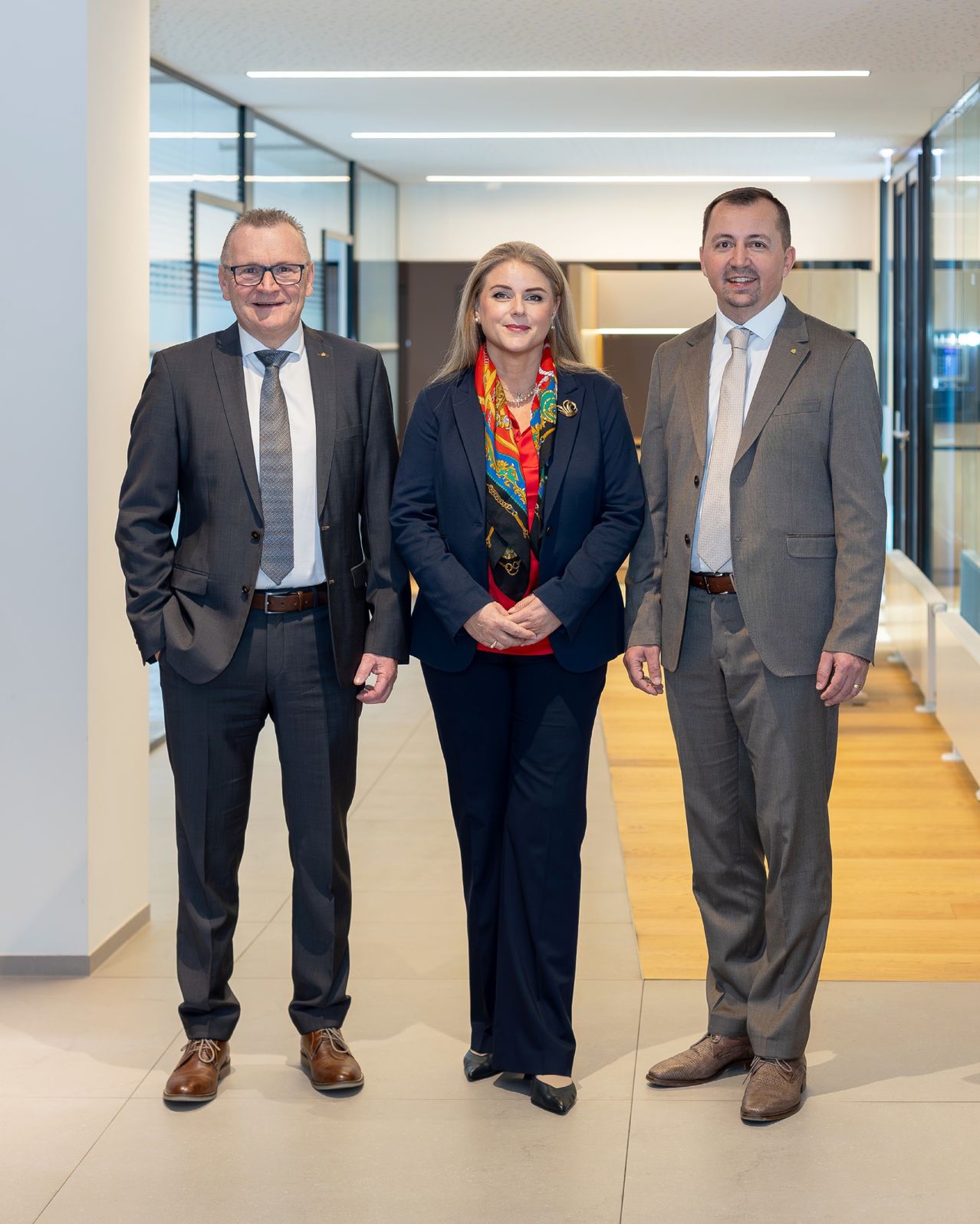 Drei formell gekleidete Personen stehen in einem modernen Büro zusammen und lächeln für ein Foto. Eine Frau in der Mitte trägt ein buntes Tuch.