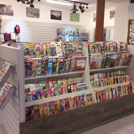 Ein Zeitschriftenregal in einem Geschäft, gefüllt mit verschiedenen Zeitschriften, einige mit Bildern von Menschen, Autos und Landschaften. Das Regal ist aus Holz und hat ein modernes Design.