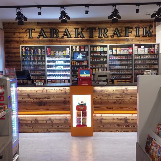 Das Innere eines Tabakladens mit einer Holzwand, einer Theke und Regalen, die mit verschiedenen Tabakprodukten bestückt sind. Der Laden hat ein modernes Design mit einer roten Theke und einer Glasvitrine.