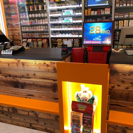 Ein Ladentresen mit Holzdesign, ein Lotterieautomat, der '28 Mio.' anzeigt, und ein Plüschtier in einem grünen Shirt.