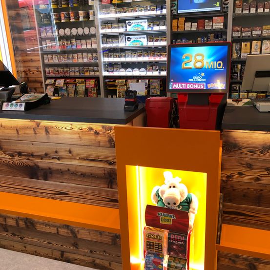 Ein Ladentresen mit Holzdesign, ein Lotterieautomat, der '28 Mio.' anzeigt, und ein Plüschtier in einem grünen Shirt.