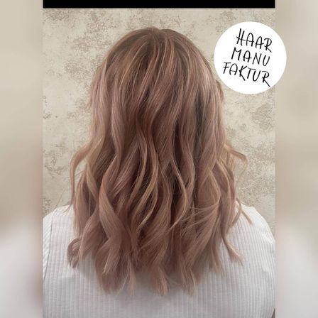 Eine Frau mit langen, welligen, roségoldenen Haaren ist mit einem weißen Oberteil gestylt. Das Bild hat einen Wasserzeichen, der 'HAAR MANU FAKTUR' in der oberen rechten Ecke sagt.