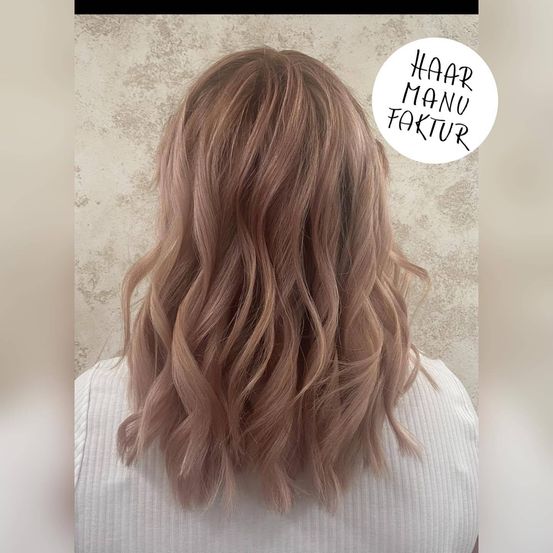 Eine Frau mit langen, welligen, roségoldenen Haaren ist mit einem weißen Oberteil gestylt. Das Bild hat einen Wasserzeichen, der 'HAAR MANU FAKTUR' in der oberen rechten Ecke sagt.
