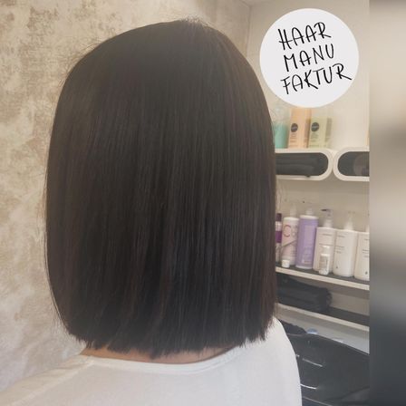 Eine Person mit glattem schwarzem Haar steht in einem Salon mit einer weißen Wand. Haarprodukte befinden sich auf dem Regal dahinter.