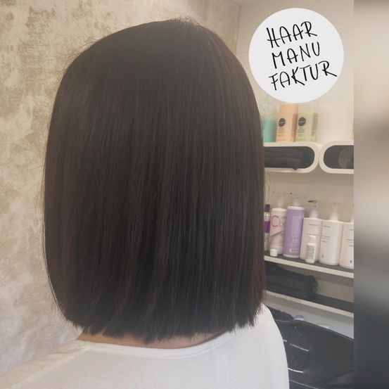 Eine Person mit glattem schwarzem Haar steht in einem Salon mit einer weißen Wand. Haarprodukte befinden sich auf dem Regal dahinter.