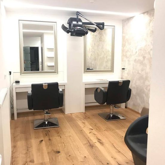 Innenraum eines Salons mit zwei schwarzen Friseurstuhlen, einem Schreibtisch und zwei Spiegeln an der Wand.