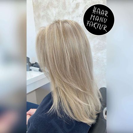 Eine Frau mit langen, glatten blonden Haaren sitzt in einem Friseurstuhl. Hinter ihr befindet sich ein weißer Schreibtisch mit einigen Gegenständen darauf und eine Wand mit einem dekorativen Kreis.