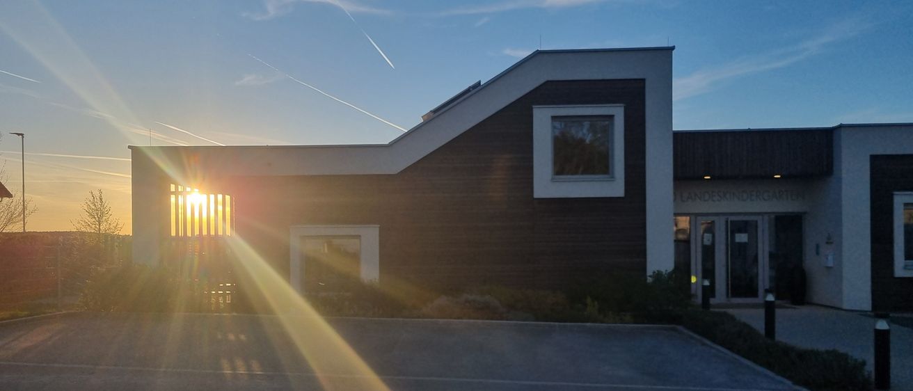 Ein modernes Gebäude mit brauner und weißer Außenseite hat ein sonnenbeschienenes Fenster und eine strahlende Sonne, die durch einen Zaun davor scheint.
