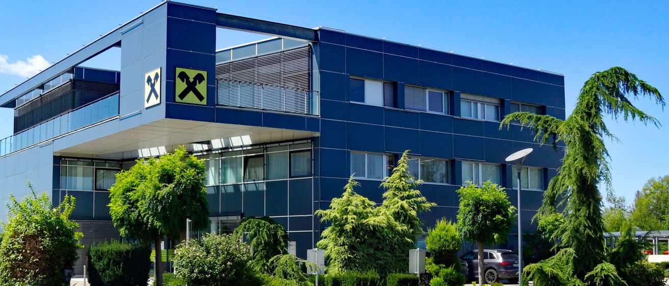 Ein modernes blaues Gebäude mit einem großen X-Logo steht unter einem klaren blauen Himmel. Es ist von verschiedenen Bäumen und Sträuchern umgeben. Ein Auto ist vorne geparkt.