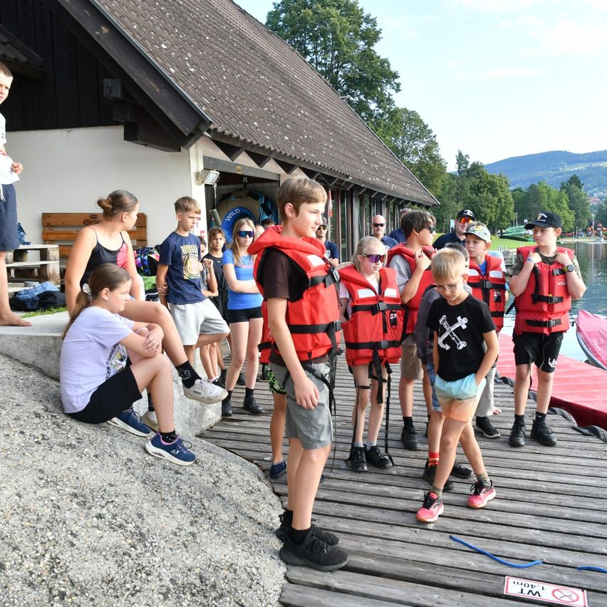 Eine Gruppe junger Menschen in Schwimmwesten steht auf einem Steg und bereitet sich möglicherweise auf eine Wasseraktivität vor. Hinter ihnen ist ein Gebäude und Bäume zu sehen, mit einer ruhigen Wasserfläche in der Ferne.