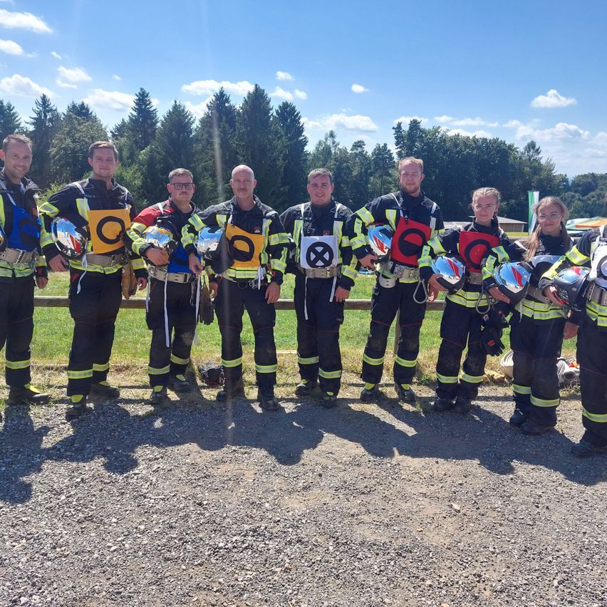 Eine Gruppe von Feuerwehrleuten in gelben und schwarzen Uniformen posiert für ein Foto und hält Helme, mit Bäumen im Hintergrund.