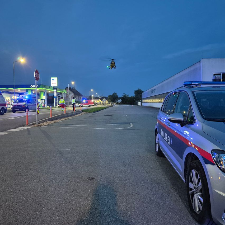 Ein Polizeiauto ist nachts am Straßenrand geparkt. Ein Krankenwagen steht an einer Tankstelle mit einem Stoppschild. Ein Hubschrauber fliegt oben.