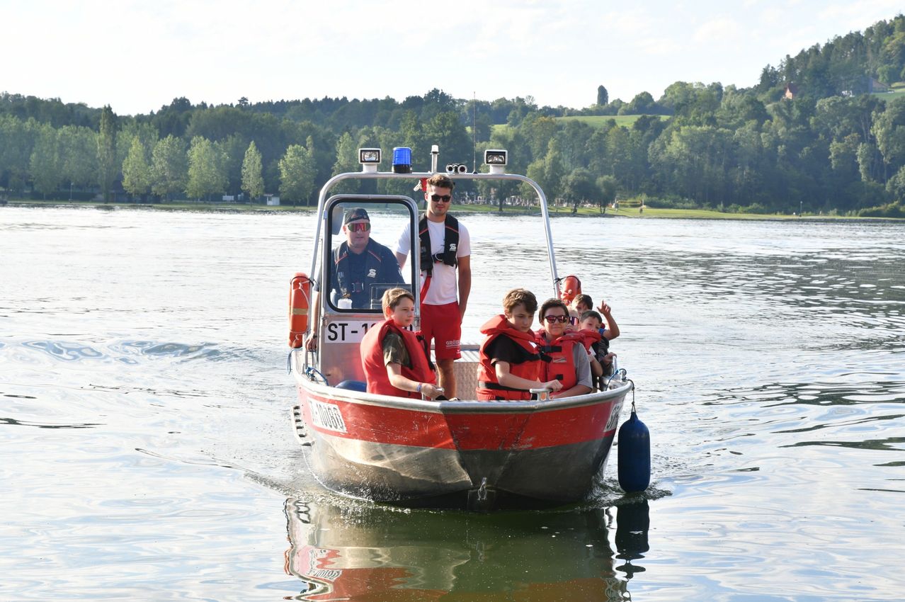 Eine Gruppe von Menschen, einschließlich Kindern, ist auf einem roten Boot mit zwei Männern in Rettungswesten. Sie befinden sich auf einem See mit Bäumen und einem Hügel im Hintergrund.