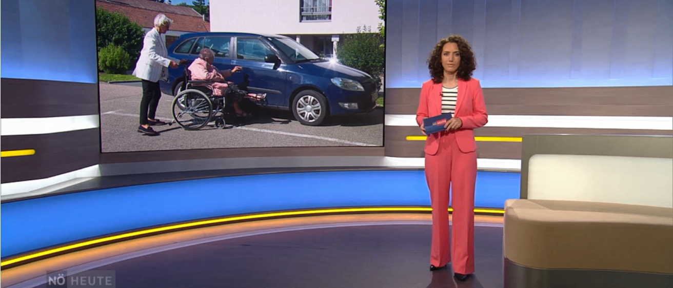 Eine Frau im Rollstuhl wird von einer älteren Frau vor einem blauen Auto unterstützt. Eine erwachsene Frau steht in der Nähe.