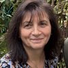 Eine Frau mit braunen Haaren, die ein gemustertes Oberteil trägt, lächelt für die Kamera im Garten.