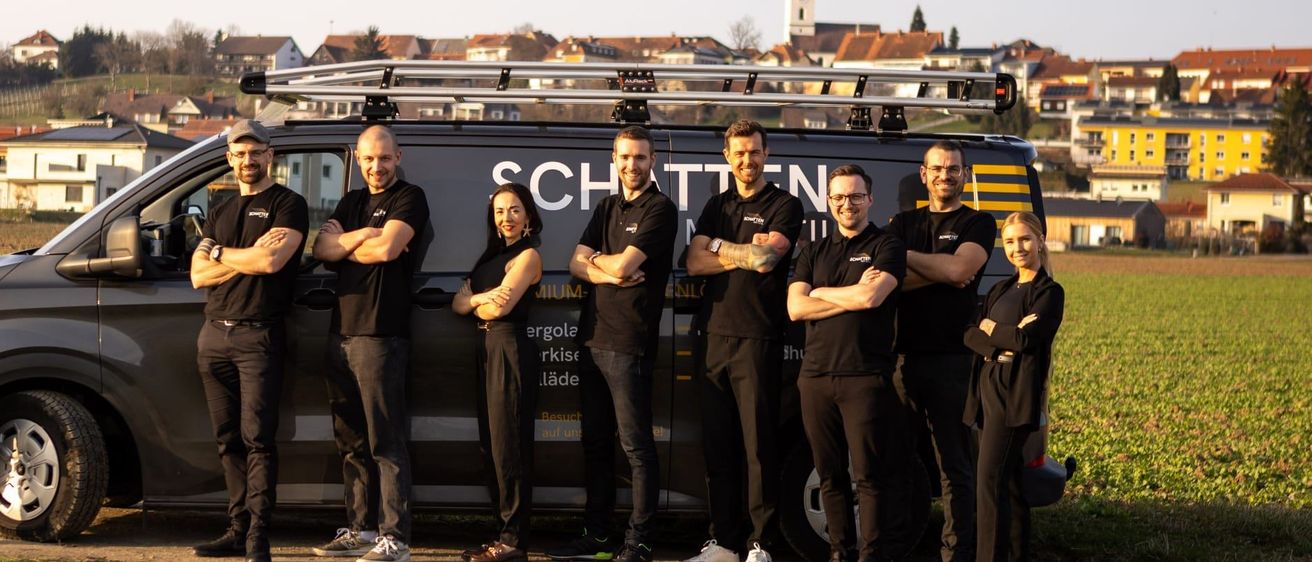 Eine Gruppe von Menschen in schwarzen Kleidern und mit Brillen posiert vor einem Van mit dem Wort 'SCHTTEN' darauf. Sie stehen auf einer Straße mit Gras und einer Stadt im Hintergrund.