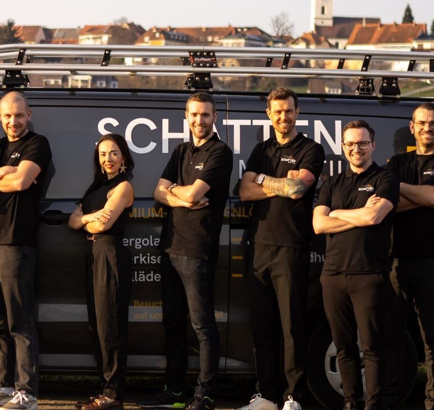 Eine Gruppe von Menschen in schwarzen Kleidern und mit Brillen posiert vor einem Van mit dem Wort 'SCHTTEN' darauf. Sie stehen auf einer Straße mit Gras und einer Stadt im Hintergrund.