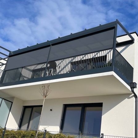 Ein Außenansicht eines modernen Gebäudes mit einem Balkon. Der Balkon hat ein schwarzes Geländer und einen versenkbaren Sonnenschutz. Das Gebäude hat große Glasfenster und einen kleinen Baum davor.