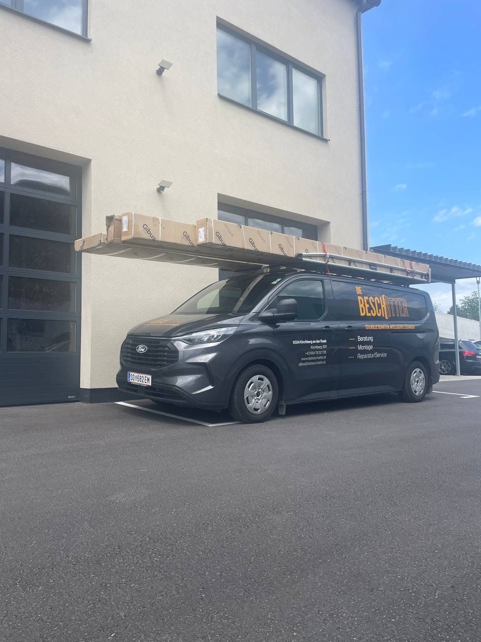 Ein schwarzer Ford-Van mit dem Text 'THE BESCHOTTER' an der Seite steht vor einem Gebäude und trägt Kisten auf dem Dach.