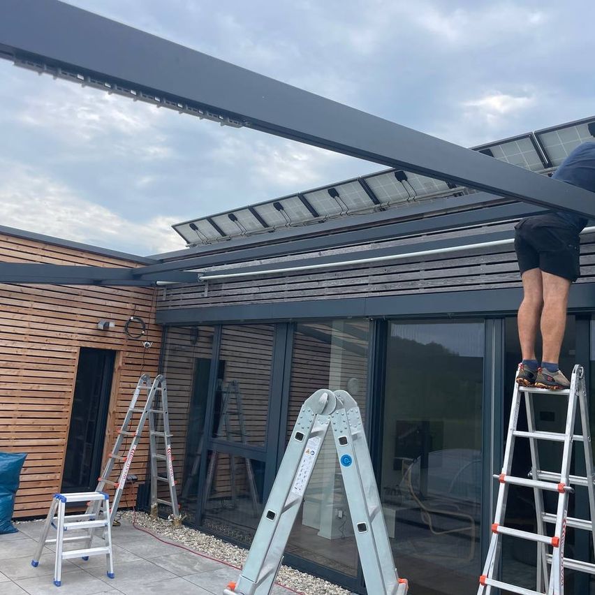Ein Mann steht auf einer Leiter unter einem solarbestückten Dach. Er arbeitet wahrscheinlich an Wartungsarbeiten. In der Nähe sind verschiedene Leitern und eine blaue Plane platziert.
