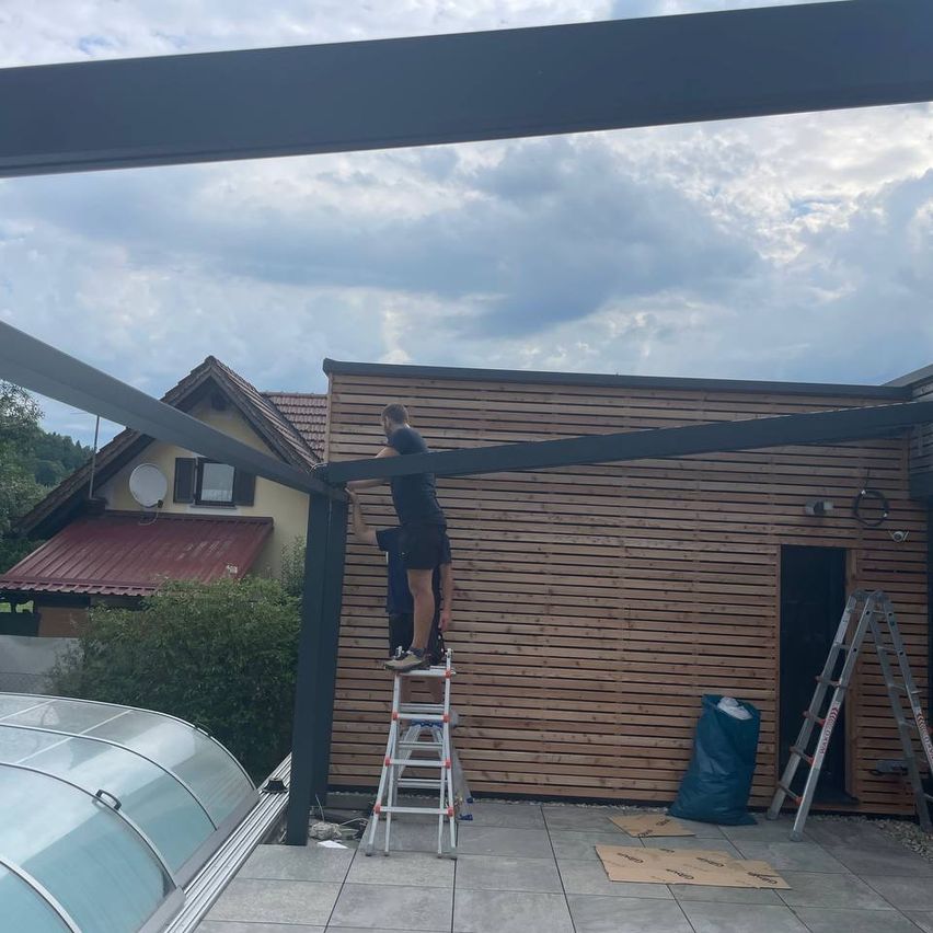 Zwei Männer arbeiten an einer Holzstruktur, möglicherweise einem Pavillon. Einer steht auf einer Leiter, während der andere auf der Struktur steht. In der Nähe befindet sich ein Haus mit einem roten Dach, einer Satellitenschüssel und einem Glasfenster. Es gibt auch Büsche, einen Müllsack und eine Leiter, die an der Wand lehnt.
