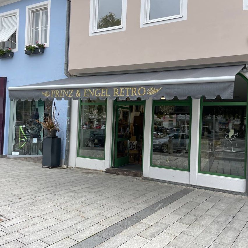 Ein Retro-Laden namens Prinz & Engel Retro mit grauem Vordach, grünen Fensterrahmen und Topfpflanzen davor.