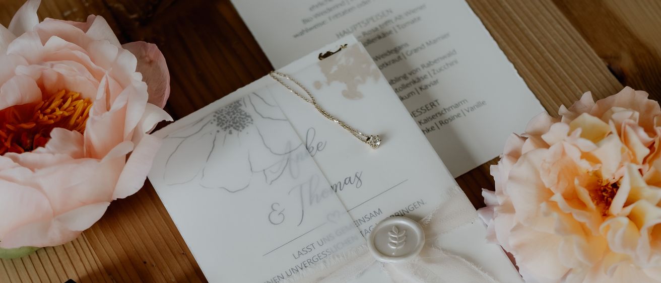 Eine Hochzeitseinladung, eine Halskette, ein Ring und eine Parfümflasche liegen auf einem Holztisch mit rosa Blumen.