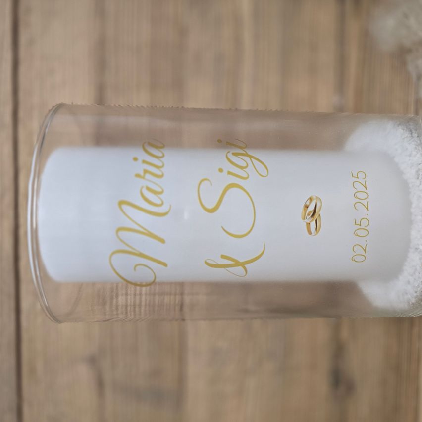 Ein zylindrischer Glasbehälter auf einer Holzoberfläche. Im Inneren des Behälters befinden sich zwei goldene Ringe und das Datum '02.05.2025' auf dem Glas geschrieben.