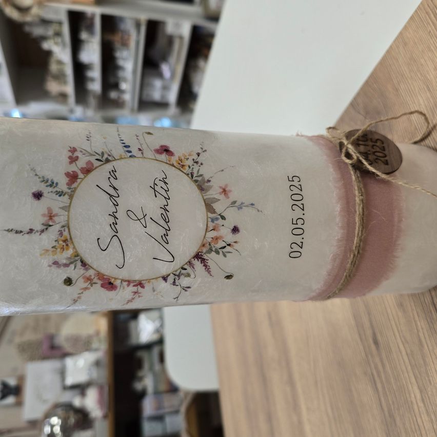Eine dekorative Kerze mit Blumendesign und einem Etikett mit dem Namen 'Sandra & Valentin'. Sie ist datiert auf den 02.05.2025 und in weißes Papier mit rosa Akzenten eingewickelt.