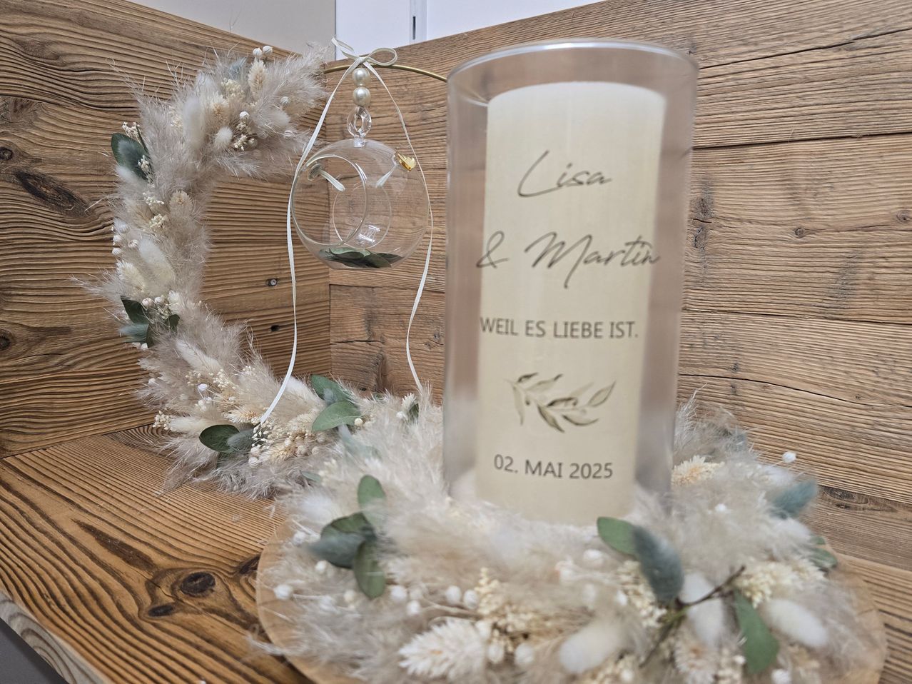 Eine Kerze auf einem Holztisch mit einer Girlande drumherum, die Kerze sagt Lisa und Martin, weil die Liebe existiert, datiert 2. Mai 2025.