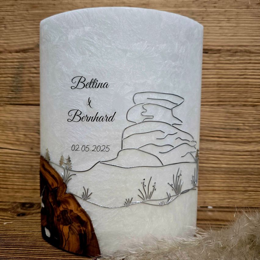 Eine weiße Kerze mit einer Landschaftsgestaltung mit Bergen und einem Baum, betitelt 'Bettina & Bernhard 02.05.2025', auf einer Holzoberfläche platziert.
