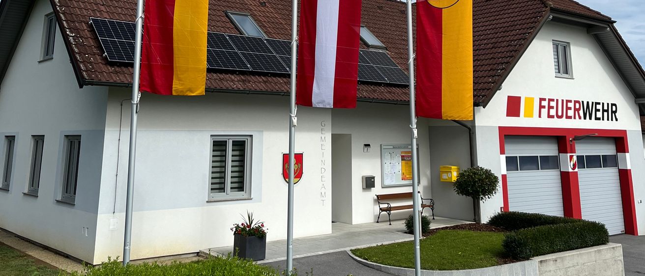 Ein weißes Gebäude mit braunem Dach, Solarpanelen und Flaggen davor. Die Flaggen sind gelb und rot mit Emblemen. Das Gebäude hat das Wort 'Feuerwehr' darauf geschrieben.