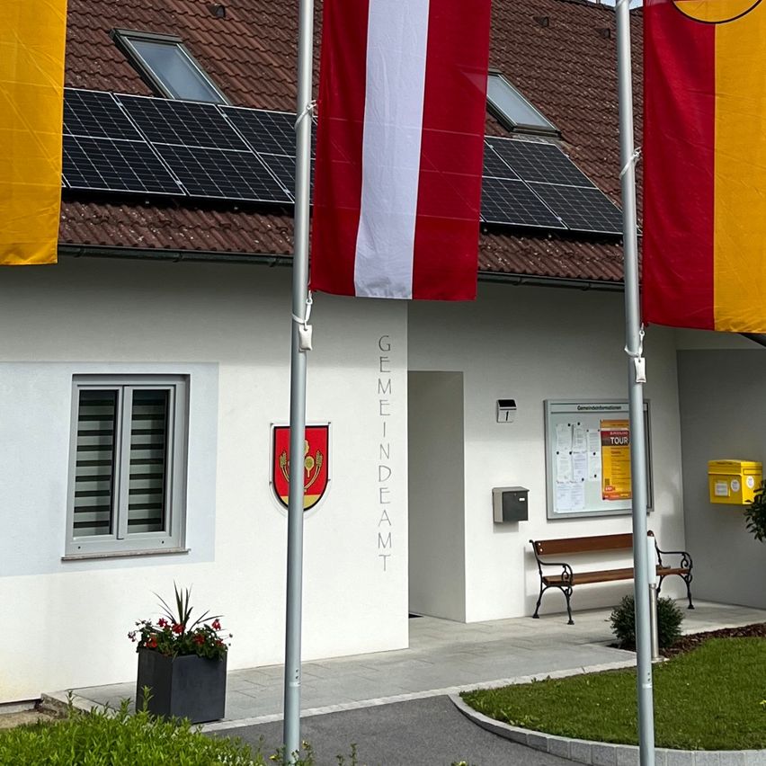 Ein weißes Gebäude mit braunem Dach, Solarpanelen und Flaggen davor. Die Flaggen sind gelb und rot mit Emblemen. Das Gebäude hat das Wort 'Feuerwehr' darauf geschrieben.