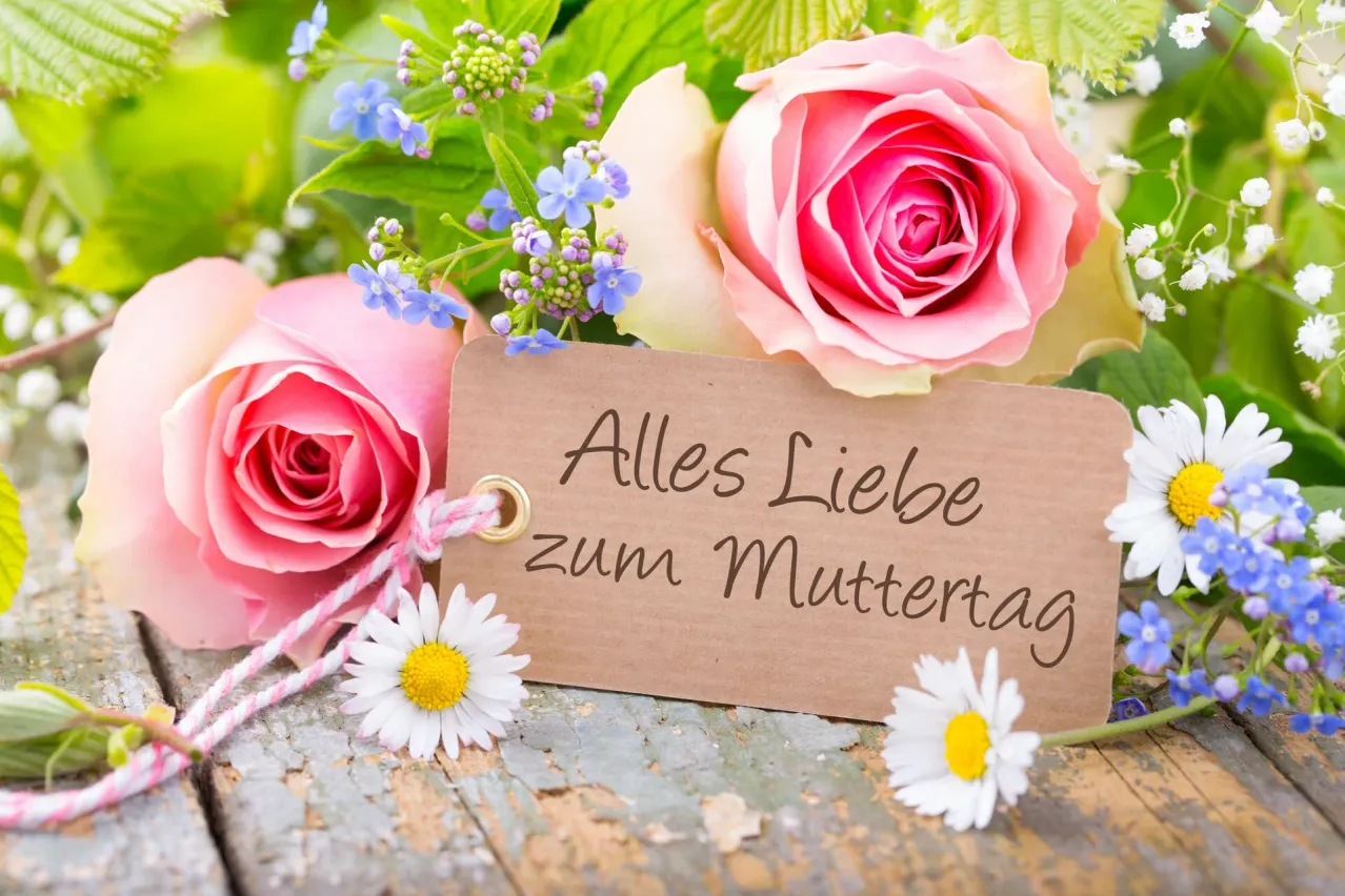 Eine Holzoberfläche hält einen Strauß pinker Rosen und blauer Blumen mit einem Schild, das Alles Liebe zum Muttertag steht.