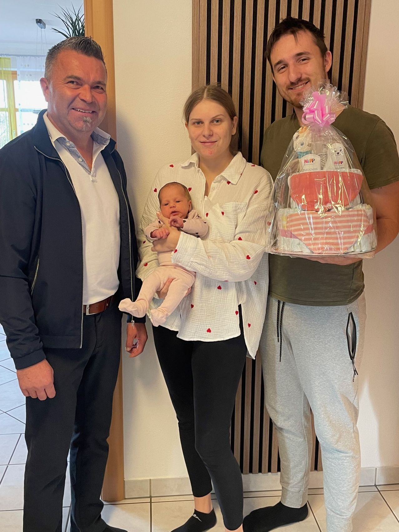 Drei Personen stehen nebeneinander. Eine Frau hält ein Neugeborenes. Ein anderer Mann hält einen Babygeschenkkorb. Der Mann auf der linken Seite trägt eine dunkle Jacke.