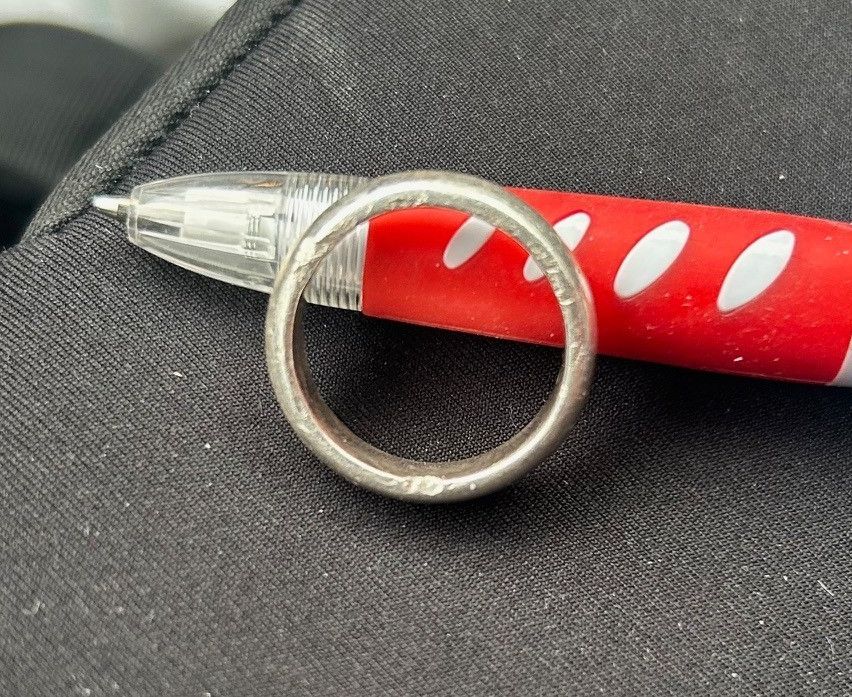 Ein silberner Ring liegt auf einem roten Stift, beide auf einer schwarzen Stoffoberfläche. Der Stift hat einen transparenten Körper mit einem roten Kappen und sichtbaren Löchern.
