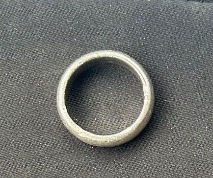Ein silberner Ring, glatt und rund, liegt auf einem dunkelgrauen Stoff. Er scheint aus Metall zu sein.