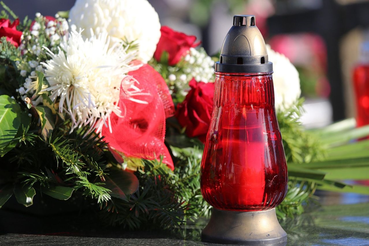 Eine rote Kerze steht in einem Glaskerzenhalter mit Metallsockel. Die Kerze ist von einem Kranz aus weißen und roten Blumen umgeben.