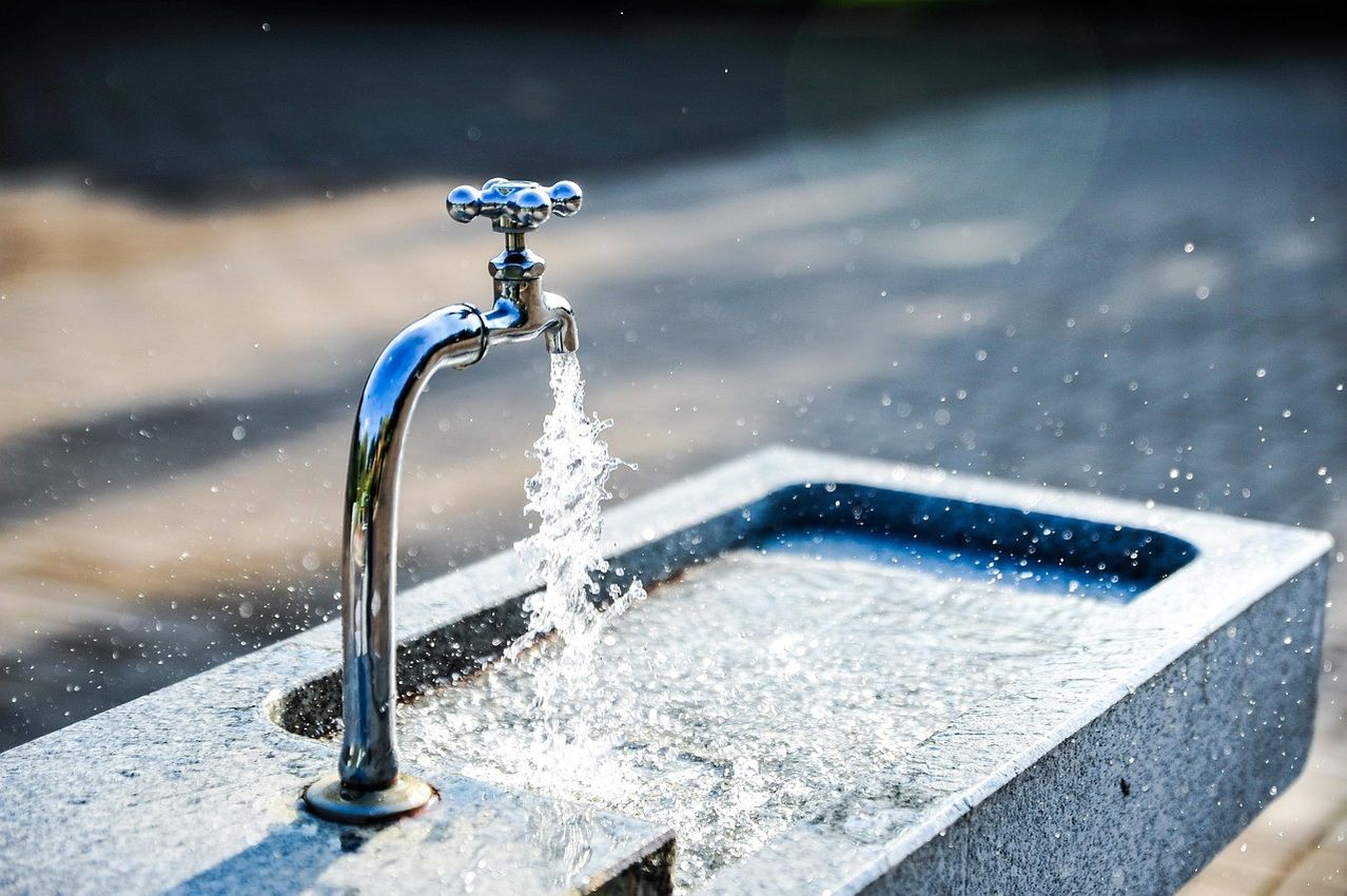 Eine Nahaufnahme eines Wasserhahns, aus dem Wasser fließt. Der Wasserhahn ist aus Edelstahl und das Wasser spritzt auf die Betonoberfläche. Der Hintergrund ist unscharf.