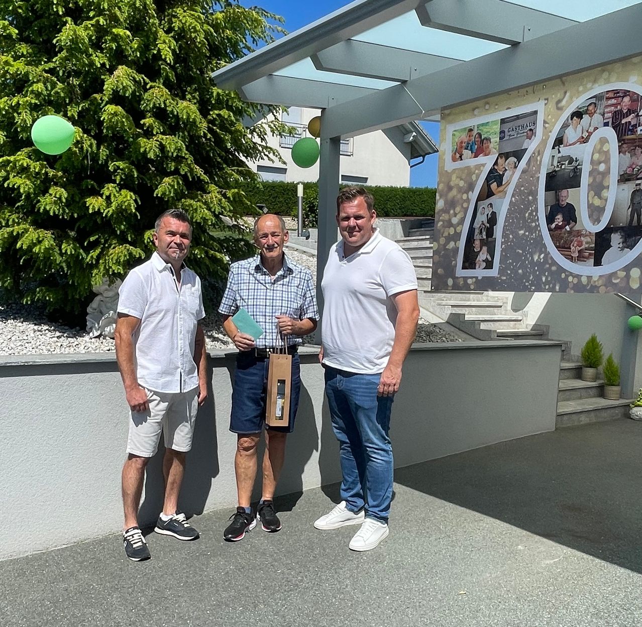 Three men stand together in front of a building with a 70th anniversary banner and green balloons. The man in the middle holds a small green envelope.