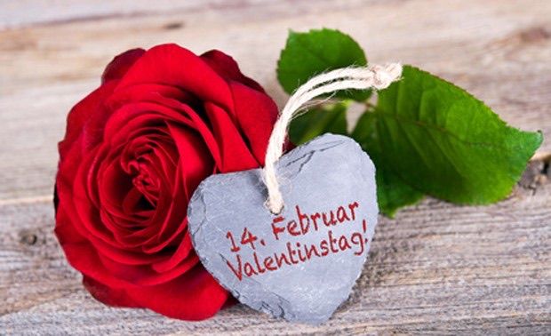 A red rose with a gray heart-shaped tag reading 14. Februar, Valentine's Day, on a wooden surface.