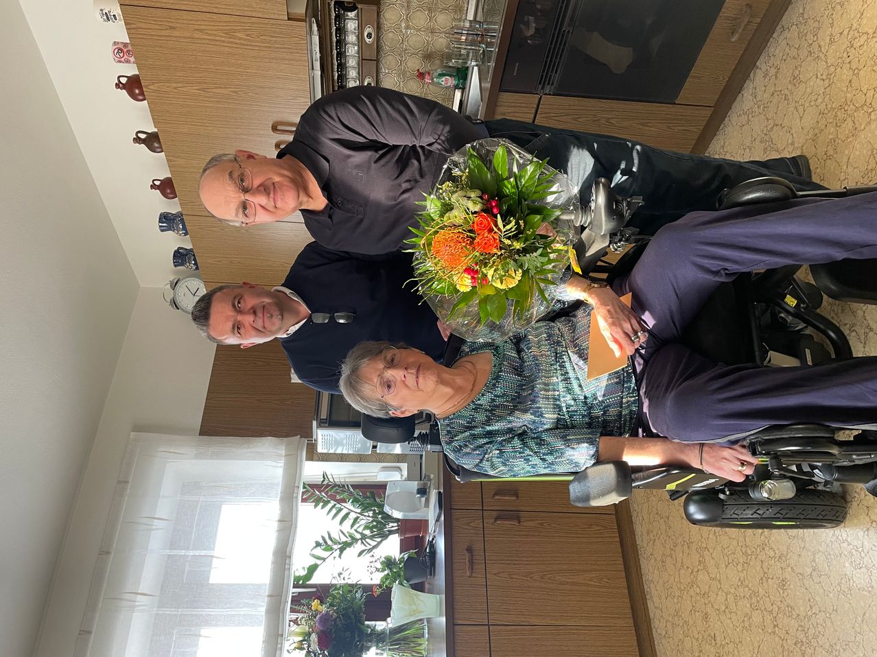 Zwei Männer posieren mit einer älteren Frau im Rollstuhl für ein Foto. Sie sind in einer Küche mit Holzschränken und Blumen auf der Arbeitsplatte.