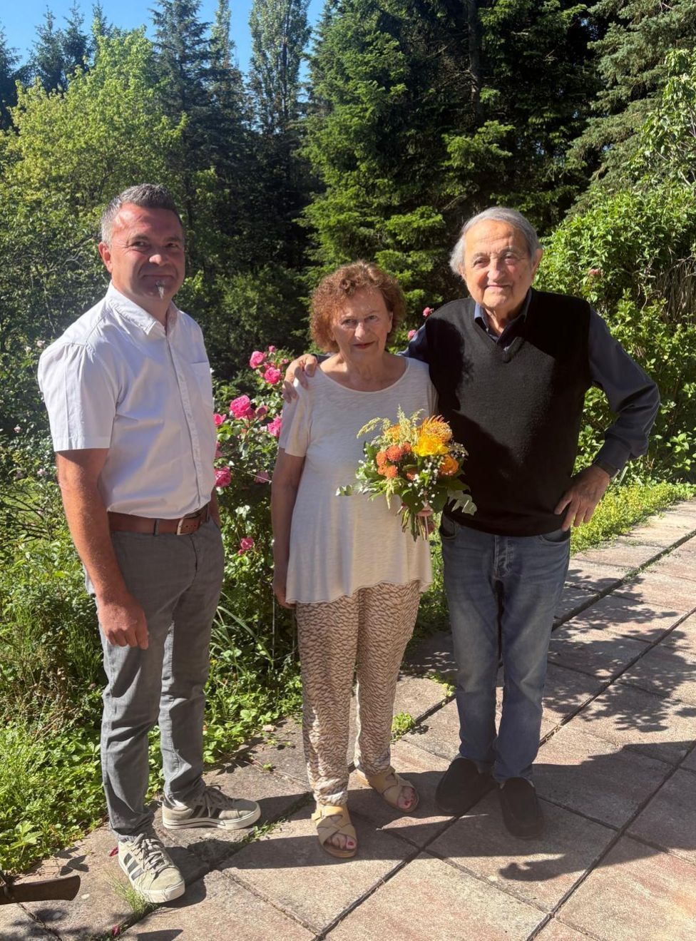Three people are standing in a garden, smiling for a photo. One woman holds a bouquet of flowers, while the man beside her has his arm around her shoulders.
