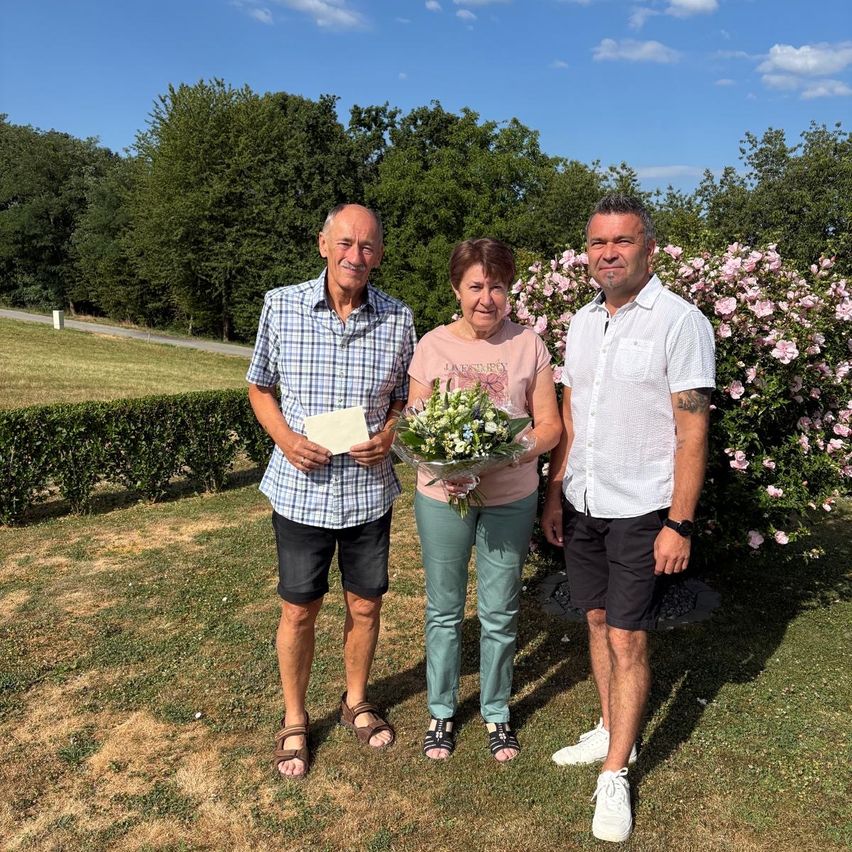 Drei Personen stehen in einem Garten mit rosa Blumen im Hintergrund, lächeln und halten einen Blumenstrauß.