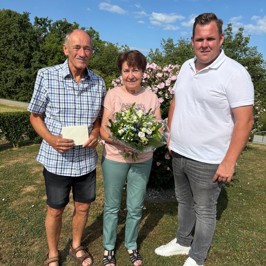 Drei Personen stehen auf einem Rasen mit Blumen im Hintergrund. Der Mann links hält ein Papier, die Frau in der Mitte hält einen Blumenstrauß und der Mann rechts trägt ein weißes Poloshirt.