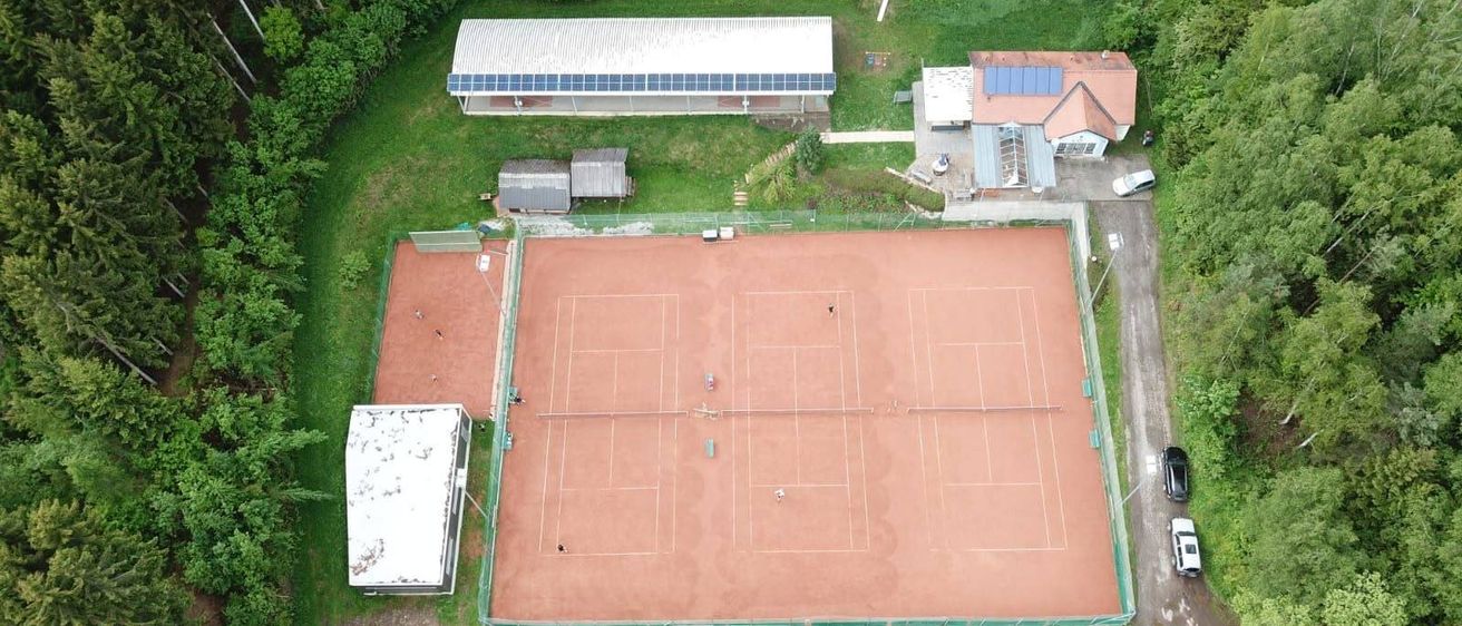 Eine Luftaufnahme eines Tennisplatzes mit Spielern auf dem Platz, umgeben von einem Zaun und Gebäuden mit Solarmodulen im Hintergrund.